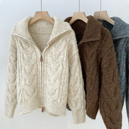 Three cable knit cardigans in beige, brown, and gray on hangers against a light background.