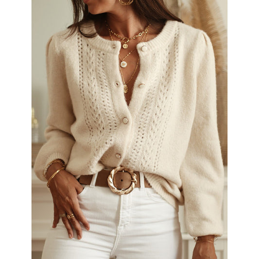 Woman wearing a cream knitted cardigan with gold jewelry and white pants.
